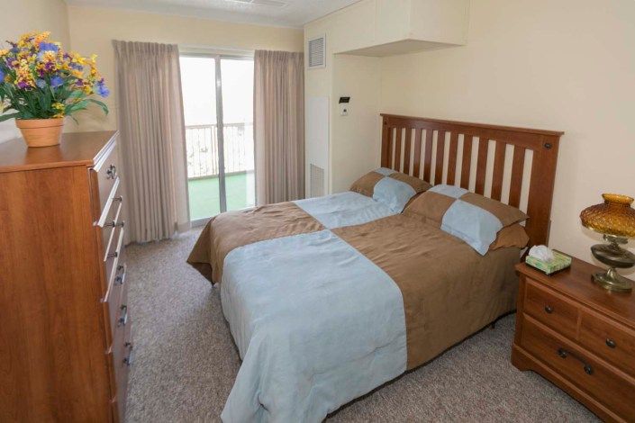 A cozy bedroom featuring a double bed with a wooden headboard, soft carpet flooring, and large windows with beige curtains.