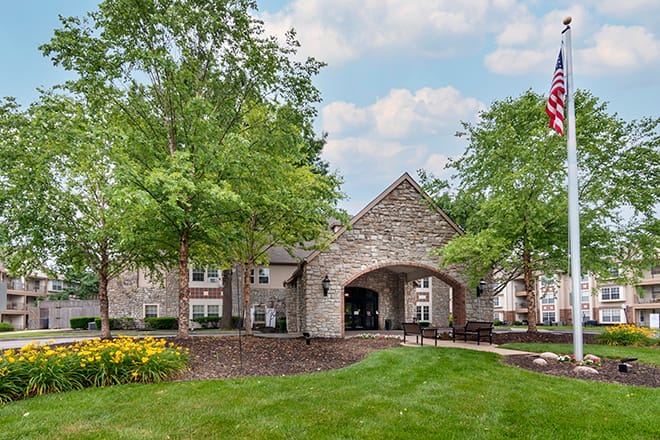 A stone building with an arched entrance surrounded by lush greenery and a well-maintained lawn under a partly cloudy sky.