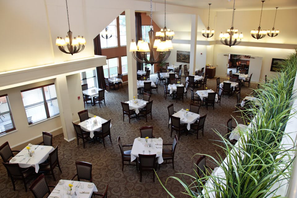 An elegant restaurant interior with neatly arranged, empty dining tables and chairs, featuring chandeliers and large windows.
