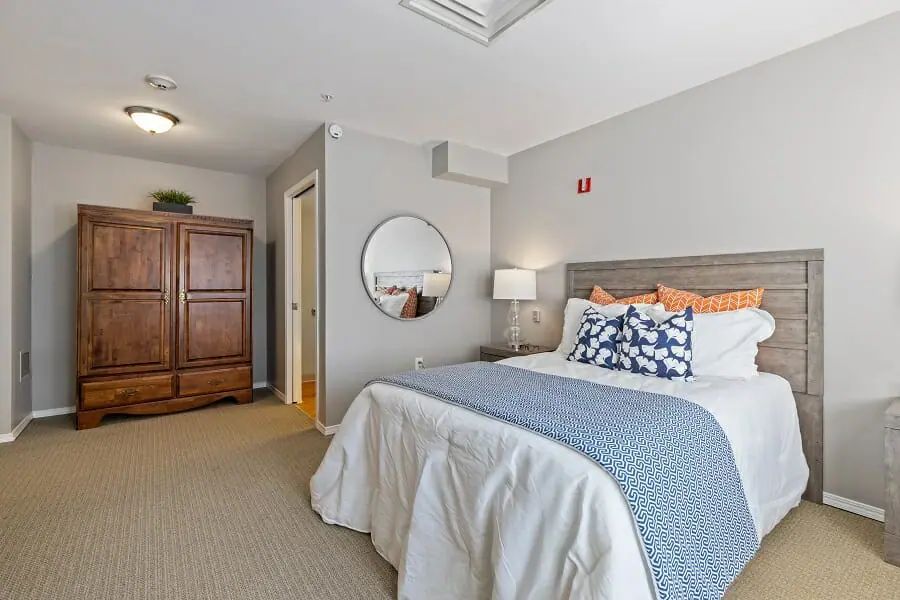 A cozy bedroom with a neatly made bed, featuring blue accents and decorative pillows. The room includes a wooden wardrobe, a round mirror, and a bedside lamp.