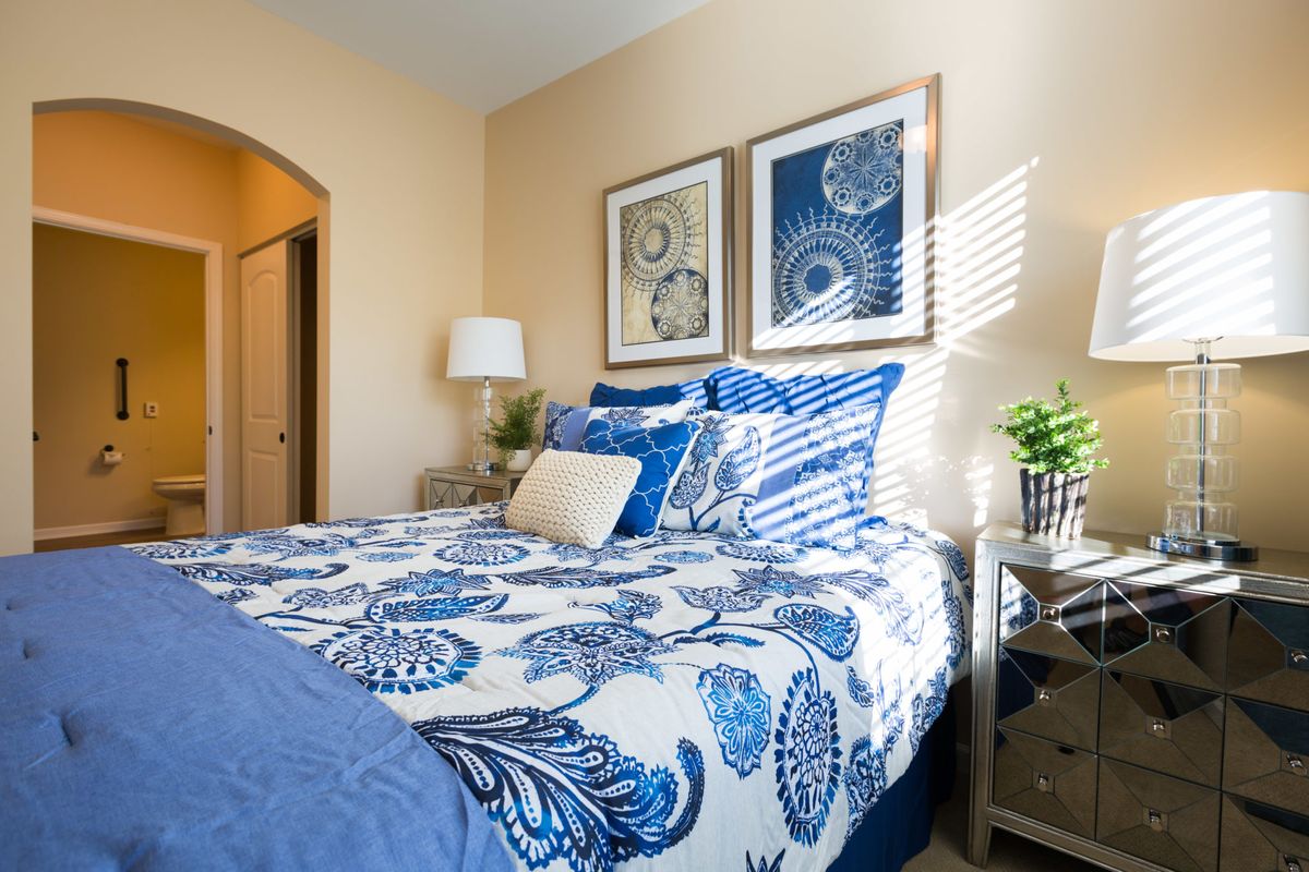 A cozy bedroom features a blue and white color scheme with ornate patterns on the bedding, complemented by framed artwork and plants.