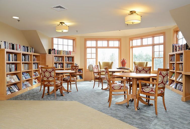 A cozy library reading room featuring wooden tables, chairs, and shelves filled with books, with large windows allowing natural light to fill the space.