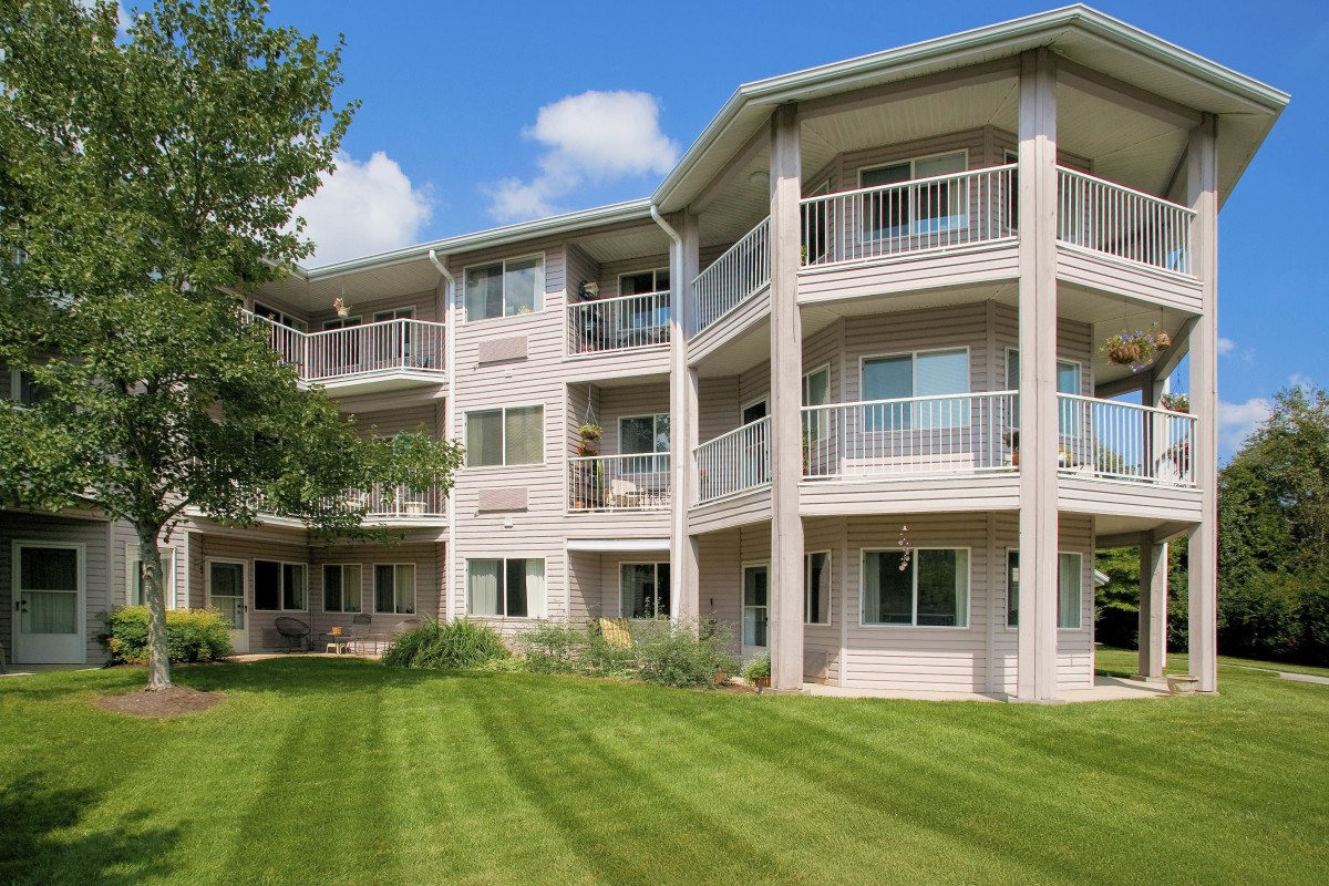A residential apartment building with multiple balconies, surrounded by a well-maintained lawn and trees under a clear blue sky.