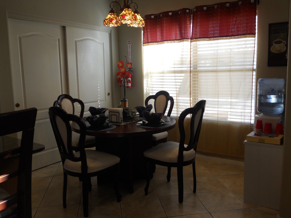 A cozy dining room with a round table and elegant chairs, illuminated by a decorative chandelier with natural light filtering through the window blinds.