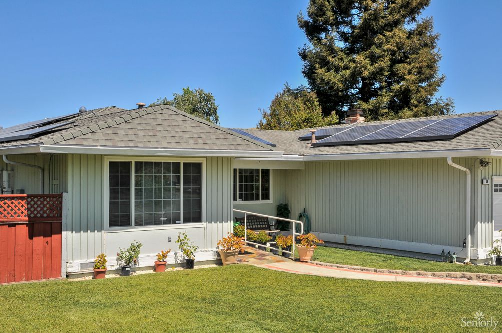 A single-story house with solar panels on the roof, surrounded by a well-maintained lawn and trees under a clear blue sky.