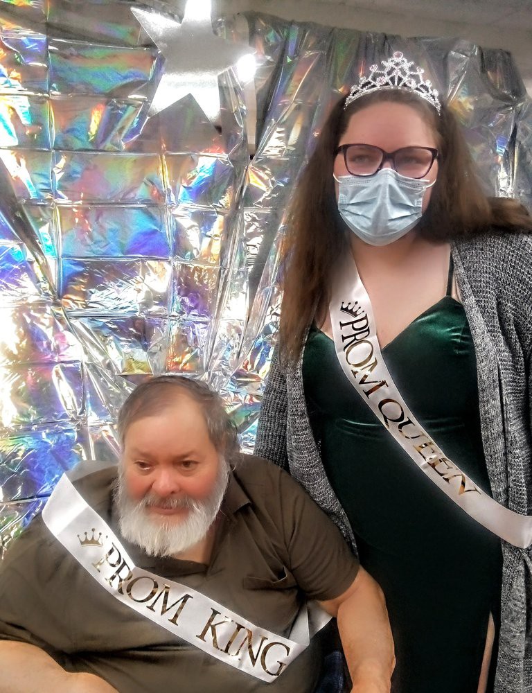 Two individuals wearing prom sashes and a tiara, posing for a photo with a shimmering background.