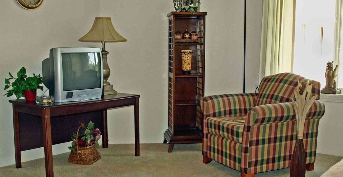 A cozy living room corner featuring a vintage TV, a plaid armchair, and a wooden cabinet with decorative items.