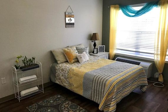 A cozy bedroom with a neatly made bed, decorative pillows, and a lamp near a window with colorful curtains.