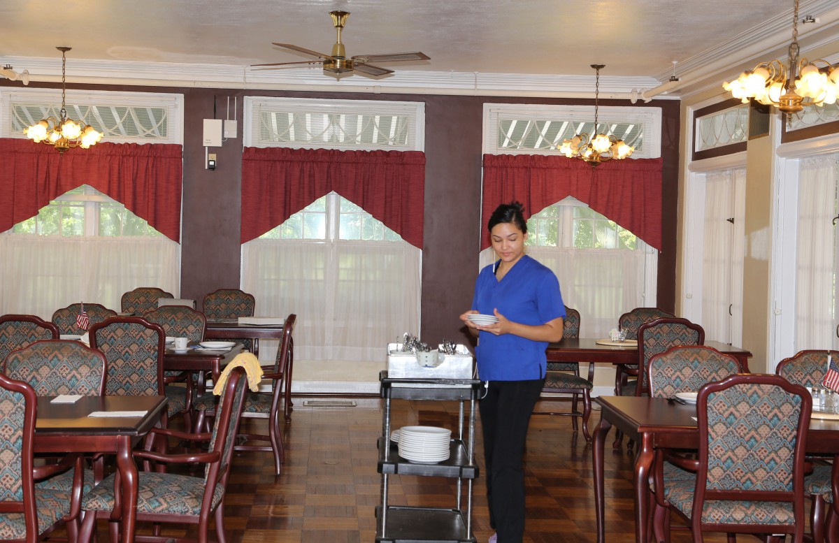 A server in a dining room setting with red curtains, pushing a cart with dishes.
