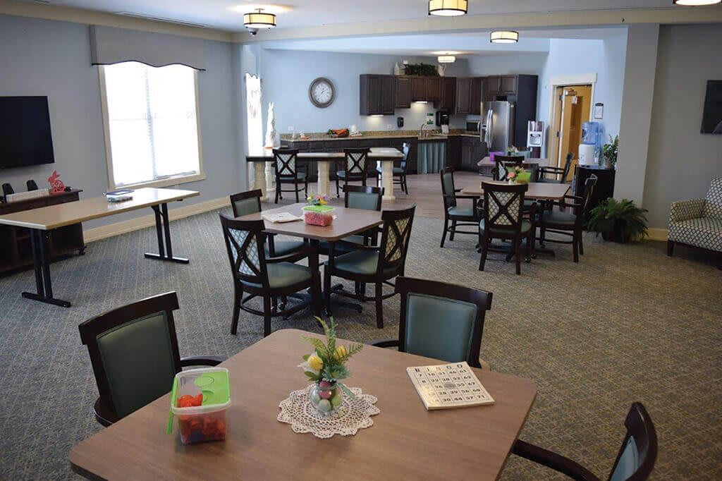 A community dining area with multiple tables and chairs, featuring a kitchen in the background.