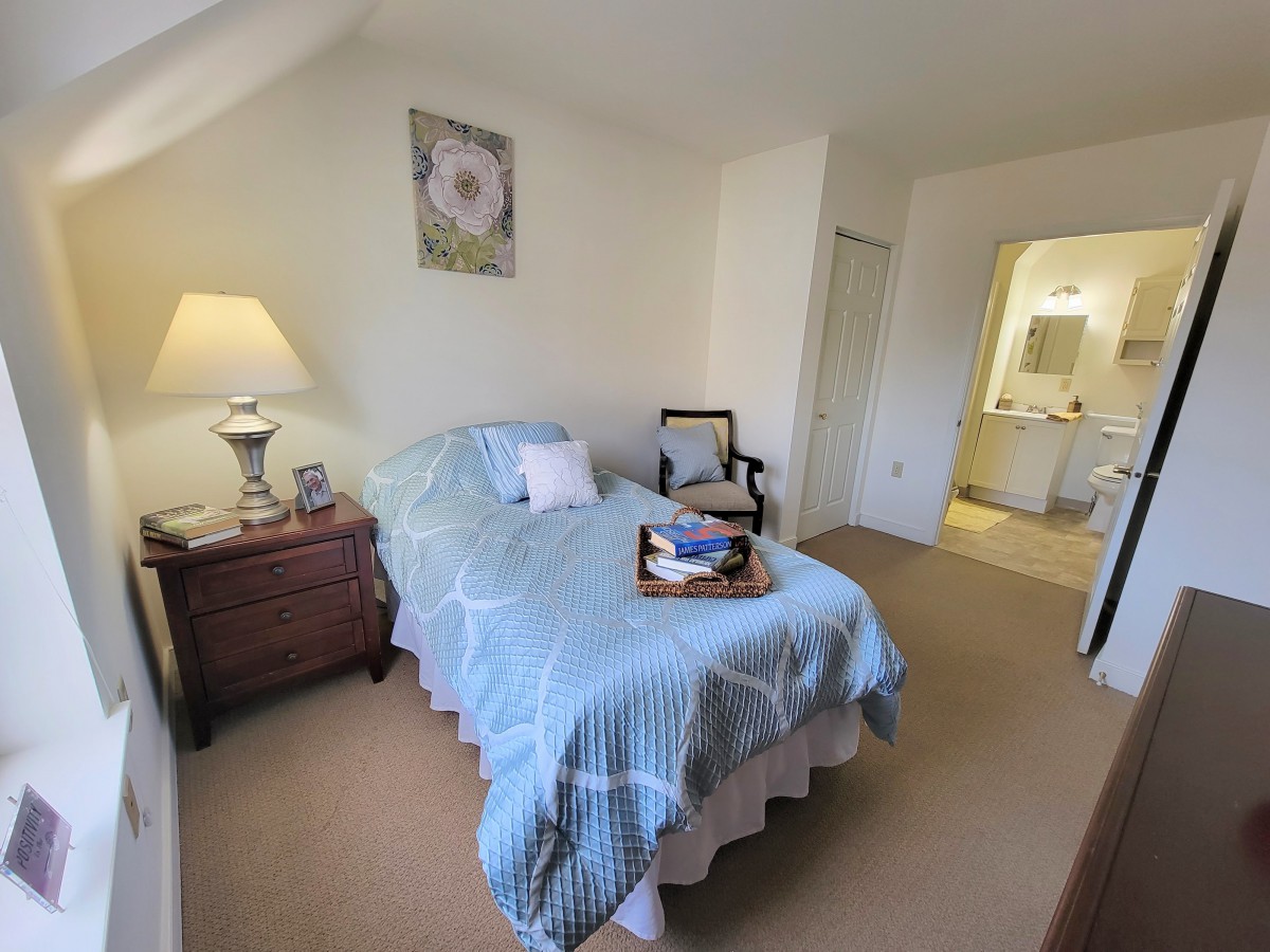 A cozy bedroom with a single bed adorned with blue bedding, accompanied by a nightstand and lamp, leading to an en suite bathroom.