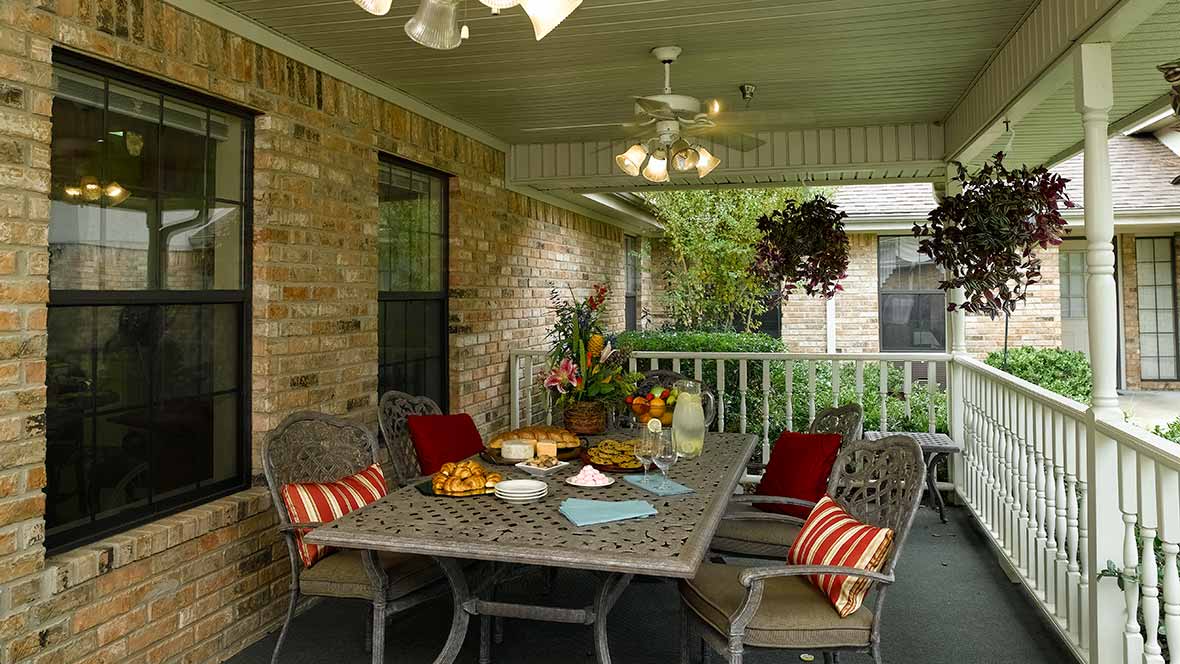 A cozy covered patio with a dining table set, decorated with cushions and surrounded by greenery.