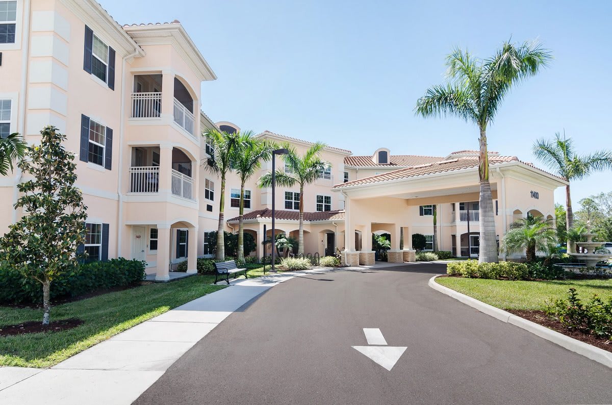 A sunny exterior view of a multi-story building with palm trees and a driveway leading to the entrance.