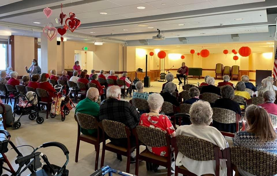 A group of seniors seated in a room decorated with red balloons and hearts, attending an event.