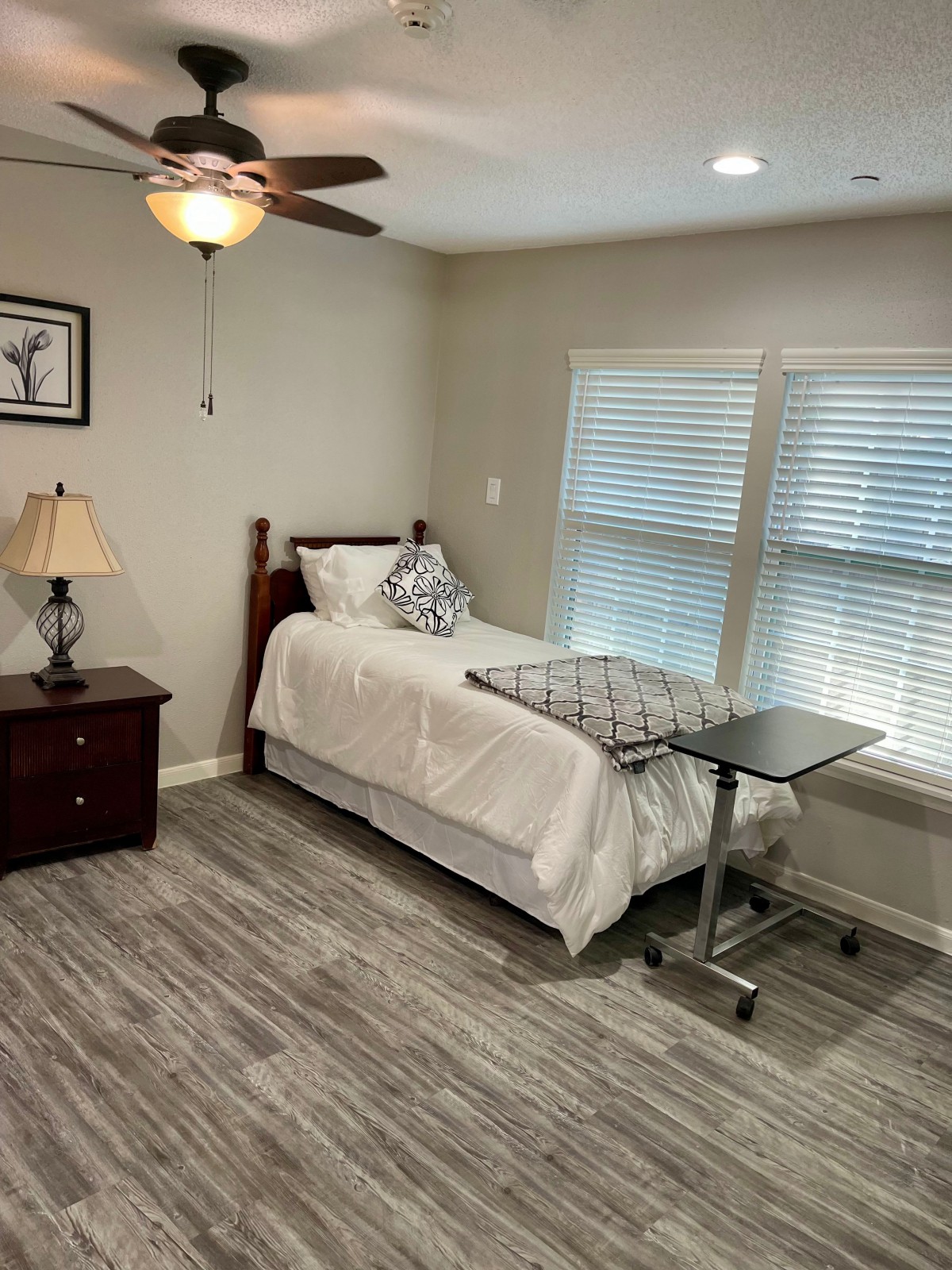 A cozy bedroom with a single bed, nightstand, and overbed table, illuminated by a ceiling fan light.