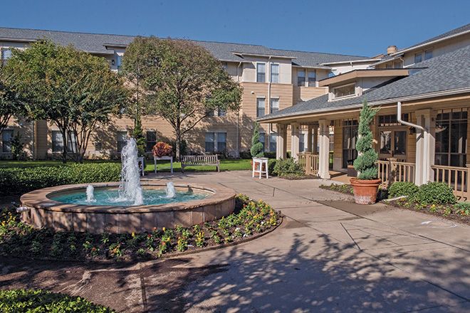 A picturesque courtyard with a central fountain, surrounded by a garden and buildings under a clear blue sky.