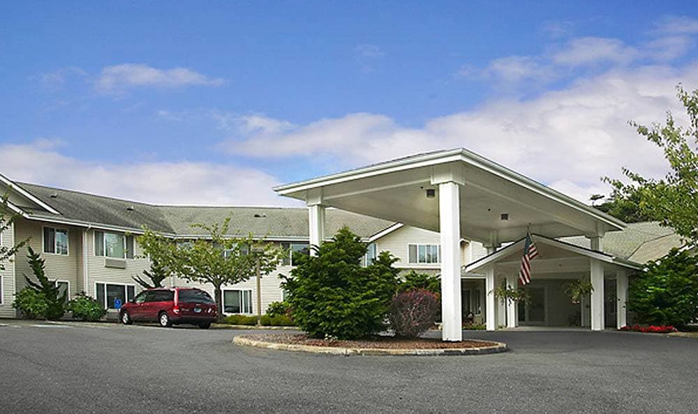 A building with a covered entrance and landscaped surroundings, including a parking area with cars.