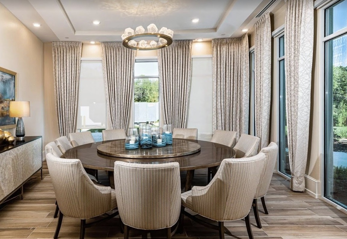 A modern dining room featuring a large round table surrounded by upholstered chairs, with elegant curtains and a decorative chandelier overhead.