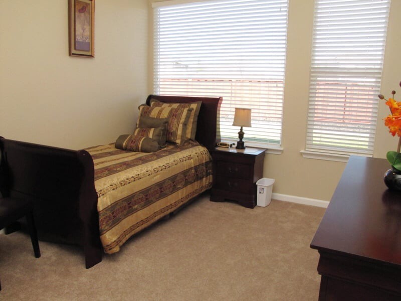 A neatly organized bedroom with a single bed, nightstand, and lamp near large windows.