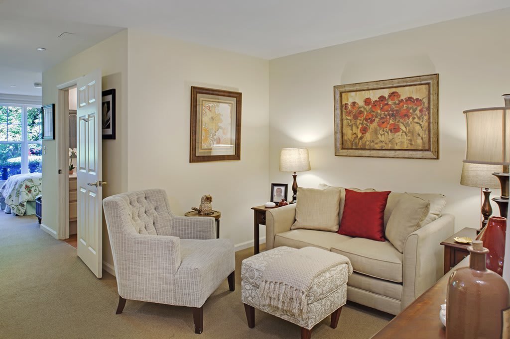 A cozy living room with a sofa, armchair, and ottoman complemented by wall art and a lamp.