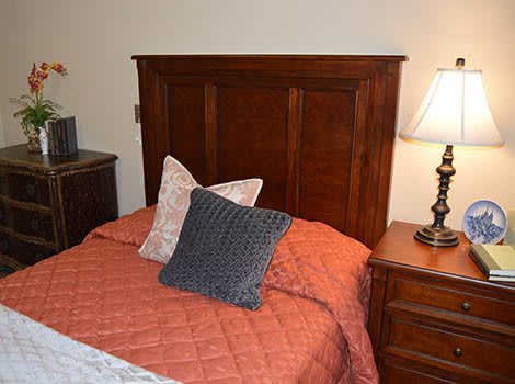 A cozy bedroom setup featuring a bed with a wooden headboard, adorned with pillows and an orange quilt, accompanied by a nightstand with a lamp.