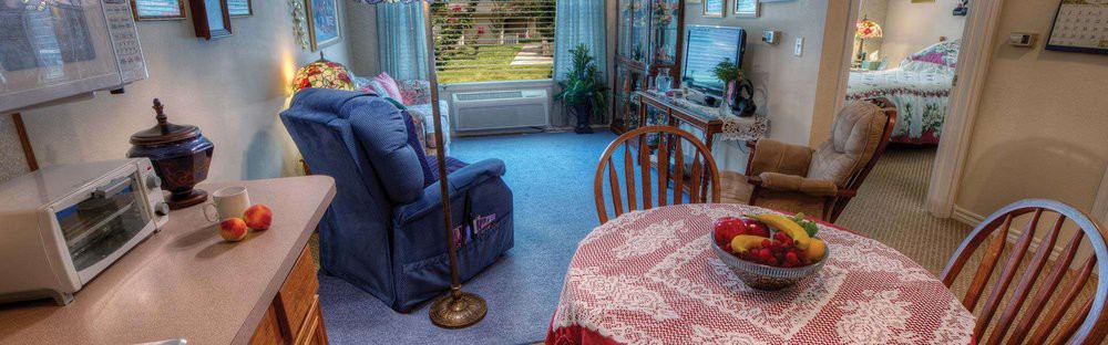 A cozy living room with blue carpet, featuring a comfortable chair, table with a patterned cloth, and large windows overlooking greenery.