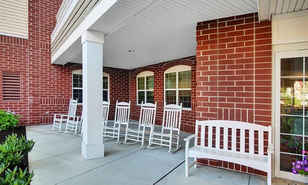 A covered porch with white rocking chairs and a bench against a brick wall.