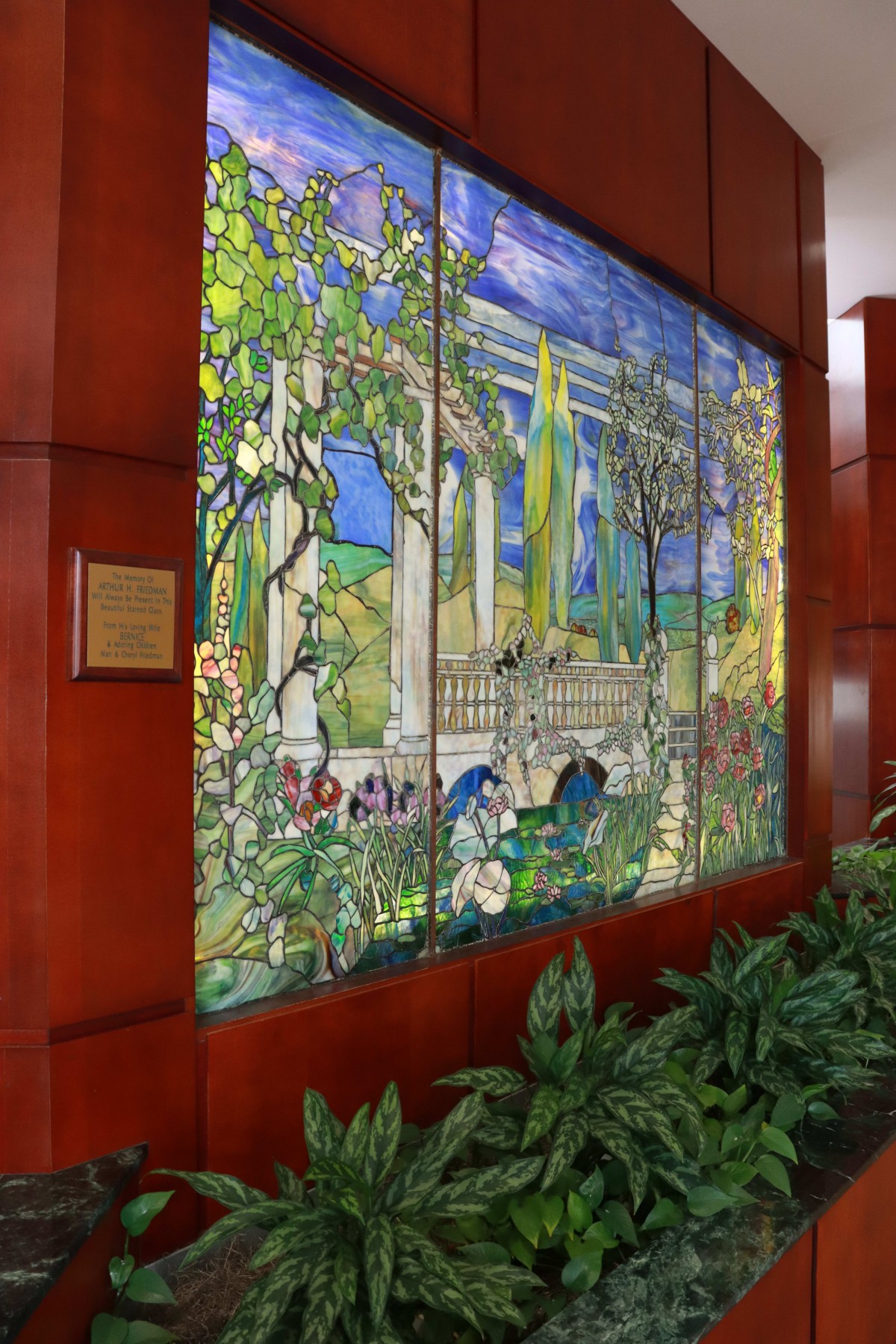 A vibrant stained glass mural depicting a serene nature scene with classical architecture, displayed indoors against a wooden wall.