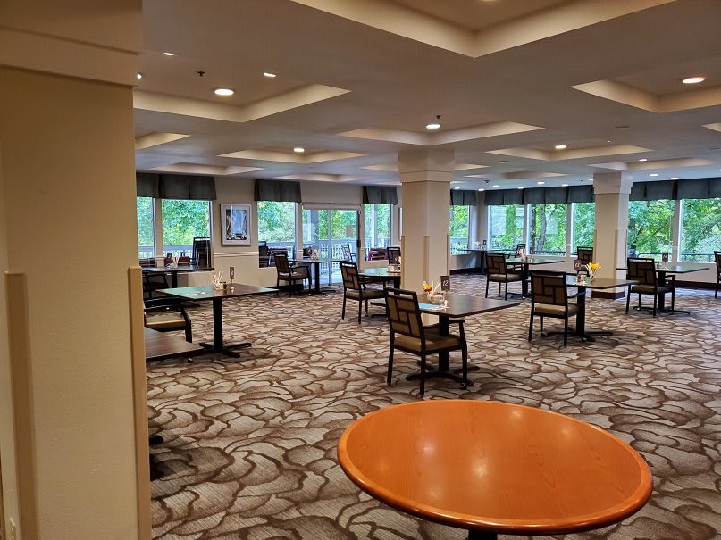 A spacious dining area with multiple tables and chairs, carpeted flooring, and large windows providing natural light.