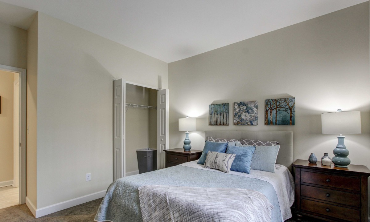 A cozy, well-decorated bedroom featuring a bed with blue and white bedding, a nightstand with a lamp, artwork on the wall, and an open closet.