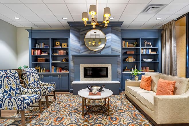 A stylish living room featuring a cozy fireplace, surrounded by bookshelves and comfortable seating, with a decorative rug and elegant chandelier.