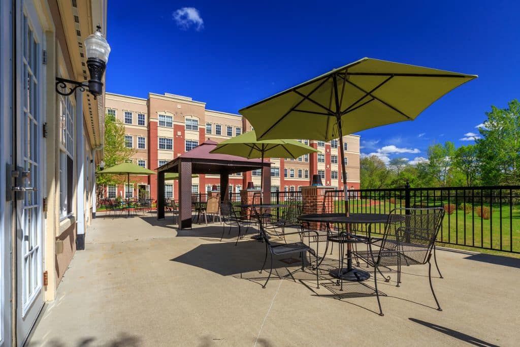 A sunny patio area with tables, chairs, and green umbrellas in front of a multi-story building.