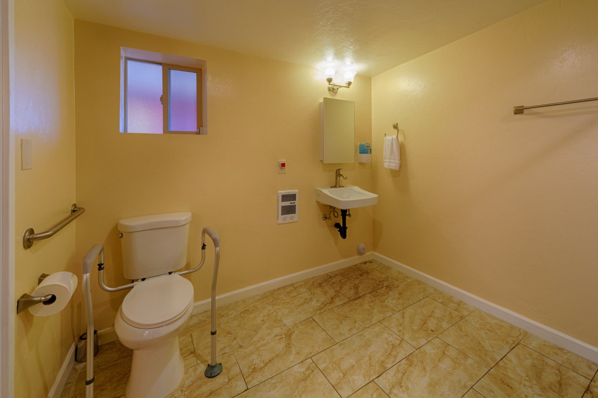 An accessible bathroom with a toilet, sink, and mirror, featuring a handrail and tiled floor.