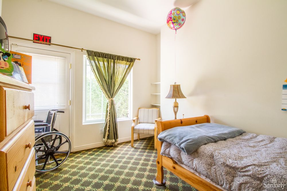 A cozy bedroom with a bed, wheelchair, and natural light coming through large windows.