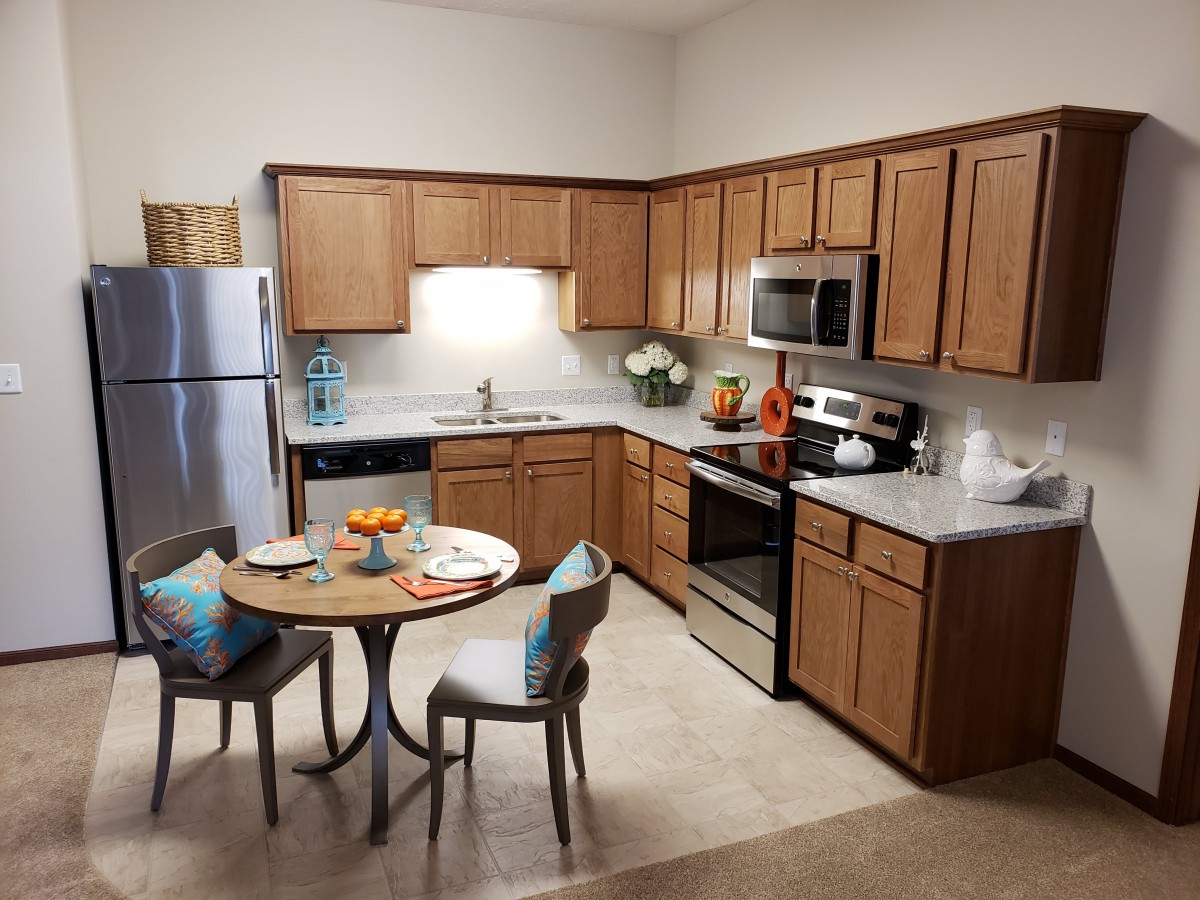 A modern kitchen with wood cabinets and stainless steel appliances, featuring a small dining table with chairs.
