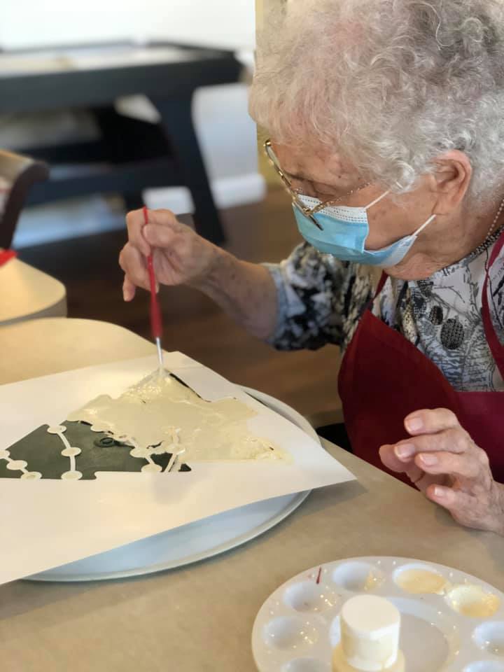 An elderly woman wearing a mask is painting using a stencil at a table.