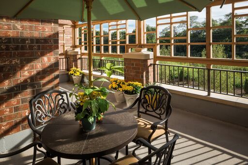 A sunny patio with elegant outdoor furniture, featuring a table under an umbrella adorned with vibrant flowers and overlooking lush greenery.
