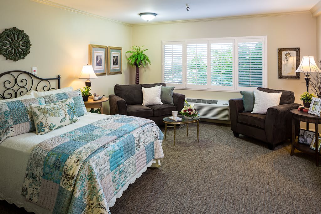 A cozy and well-decorated bedroom featuring a bed with a floral quilt, two sofas, and large windows allowing natural light.