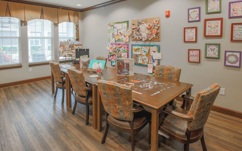 A cozy dining area with a large wooden table and upholstered chairs, adorned with colorful artwork and natural light from the windows.