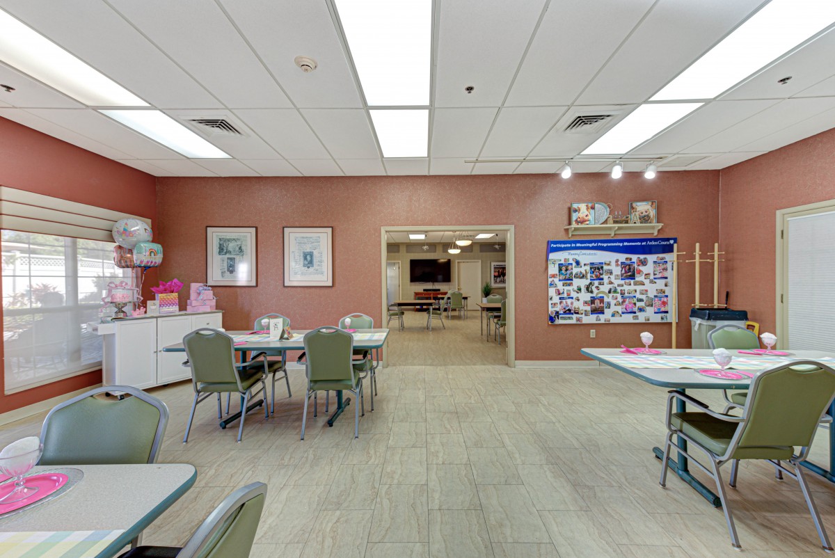 A spacious community center room equipped with tables and chairs, featuring a bulletin board and ceiling lights.