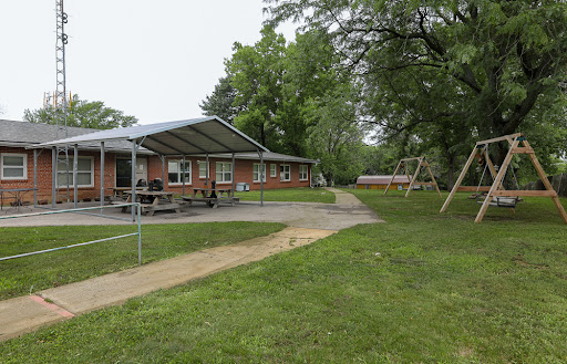 A view of a recreational building surrounded by green grass and trees, with a playground visible in the background.