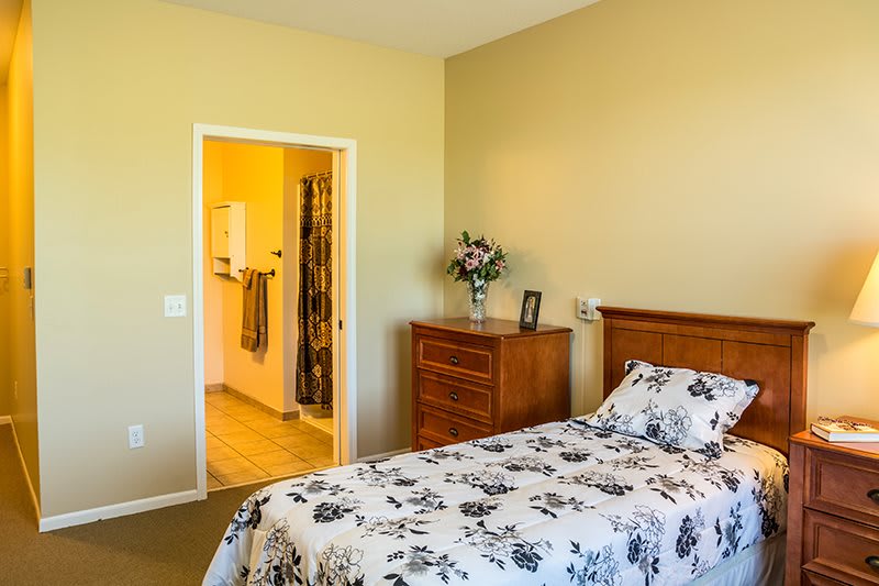 A cozy bedroom featuring a bed with floral bedding, a wooden dresser, and an open doorway to a bathroom.