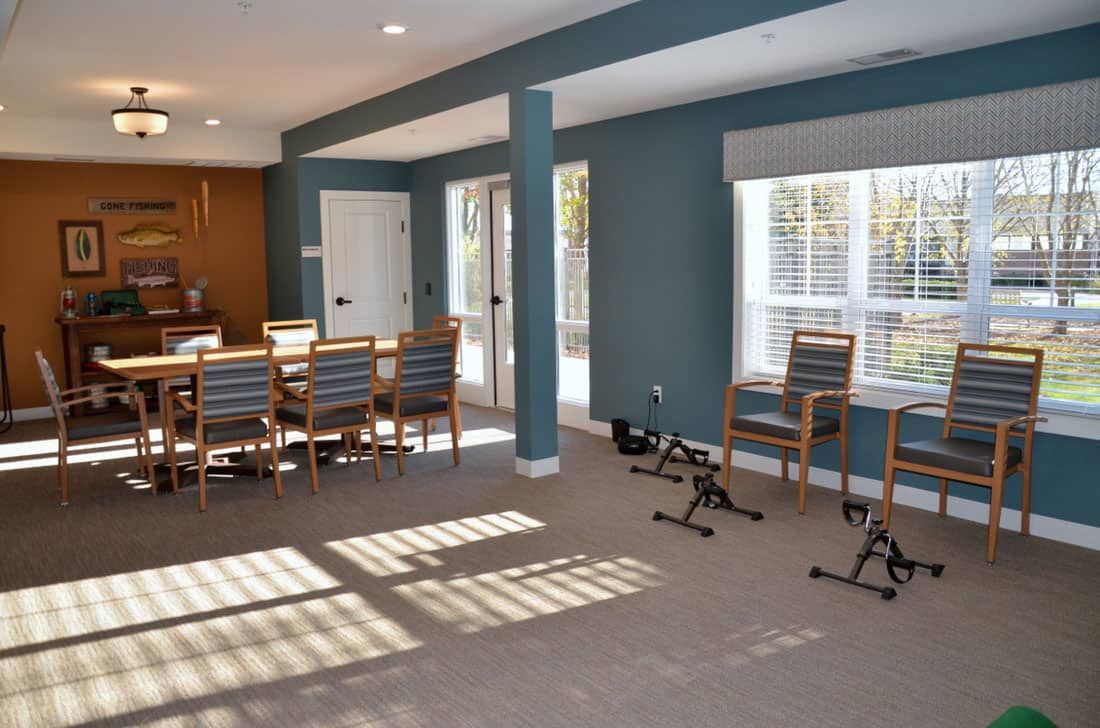 A well-lit room featuring a dining area with a long table and chairs, along with exercise equipment near large windows.