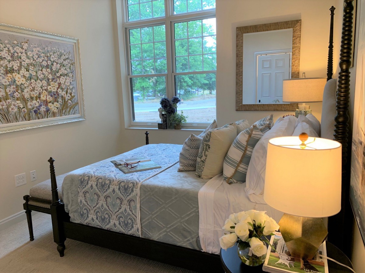 A cozy, well-decorated bedroom featuring a bed with multiple pillows, a lamp on the side table, artwork on the wall, and a window providing natural light.