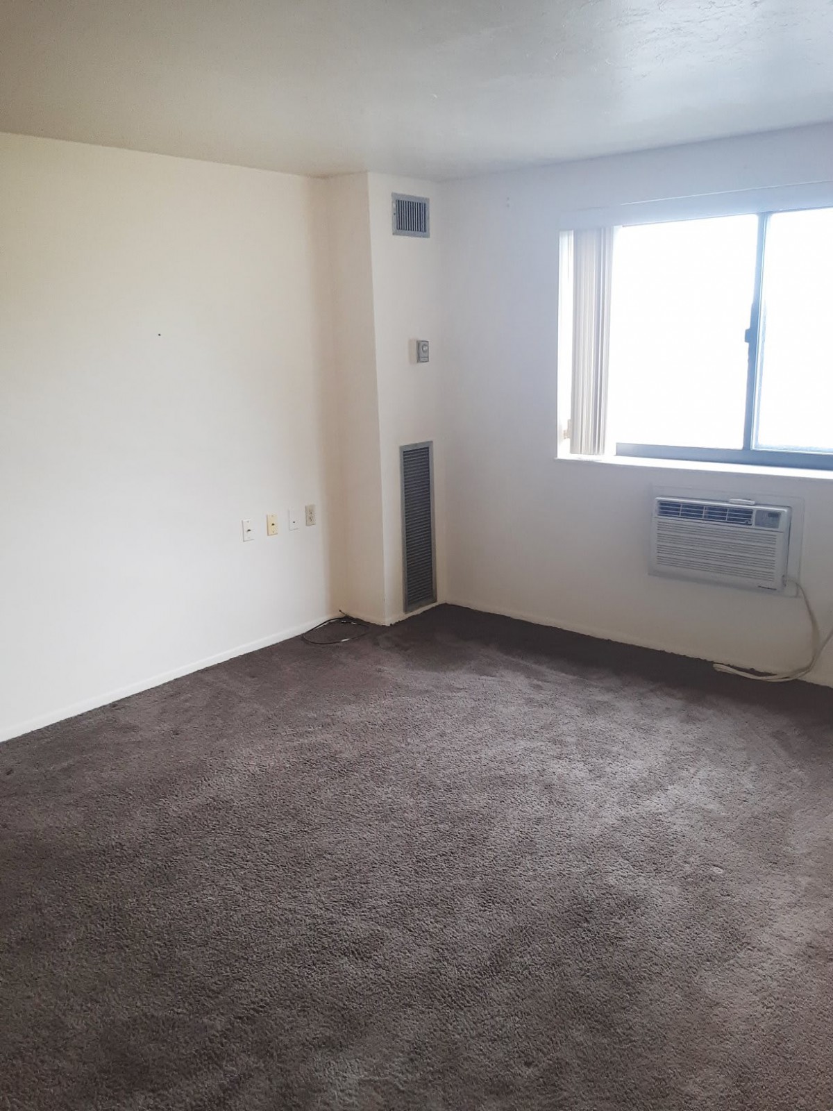 An empty room with carpet flooring, a window, and an air conditioning unit.