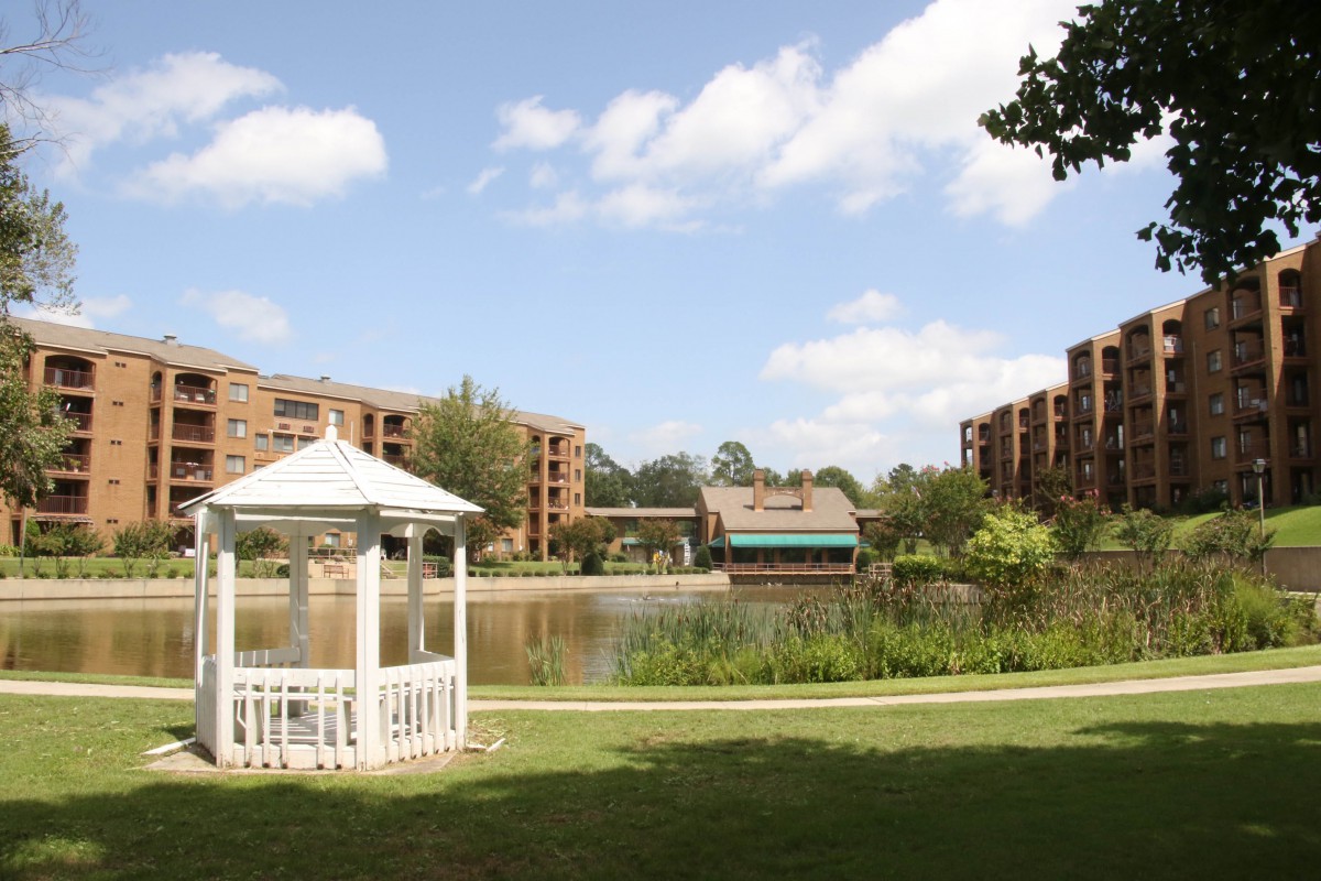 A scenic view of a park with a gazebo, lake, and surrounding apartment buildings under a partly cloudy sky.