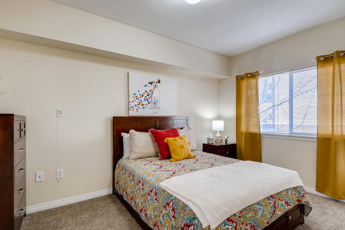 A cozy bedroom with a neatly made bed, colorful pillows, and wall art next to a window with curtains.