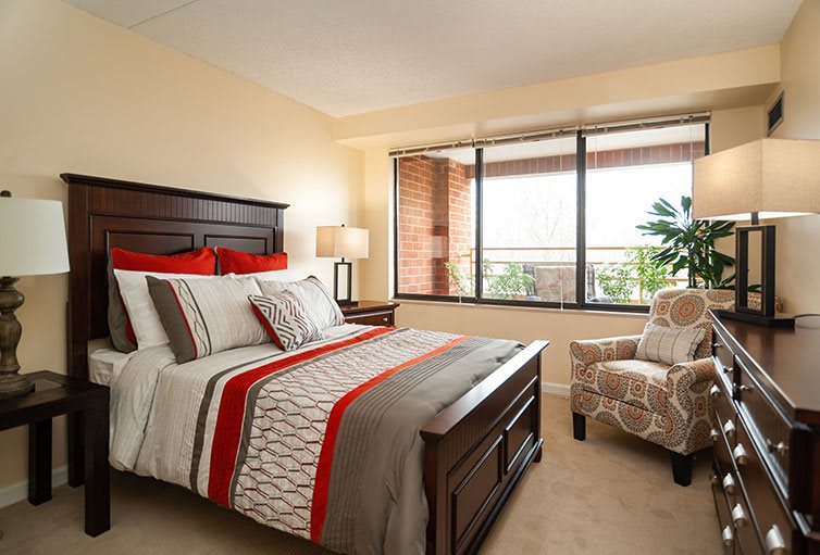 A cozy bedroom with a neatly made bed, featuring red and grey bedding, a large window, and an upholstered chair.