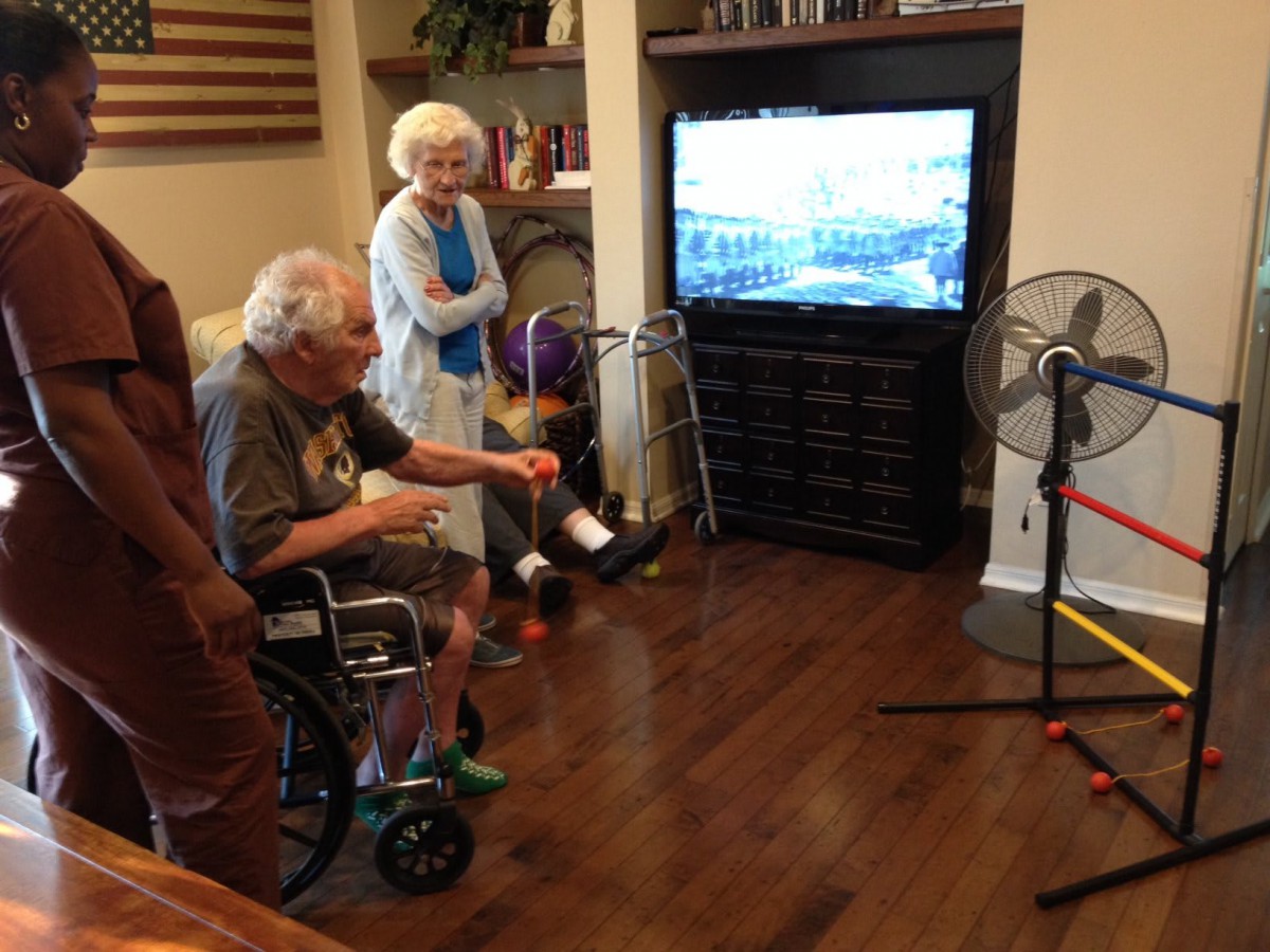 An elderly man in a wheelchair and an elderly woman are watching TV in a cozy living room.