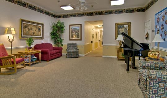 A cozy living room with a red couch, a gray chair, and a piano, leading to a hallway.